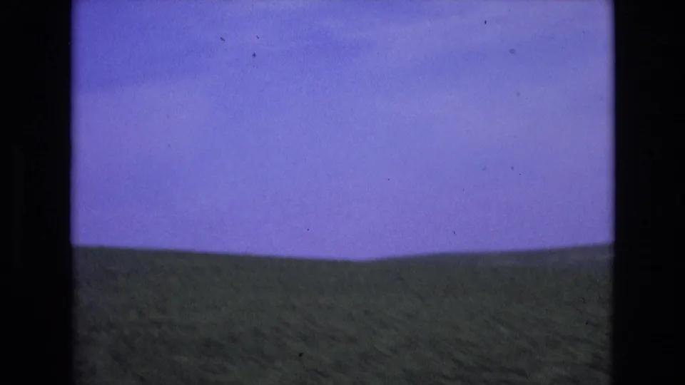 Metadata-supported 1972 Spain home-movie frame showing a green valley and low hills beneath a saturated blue sky, framed by dark 8mm gate edges.