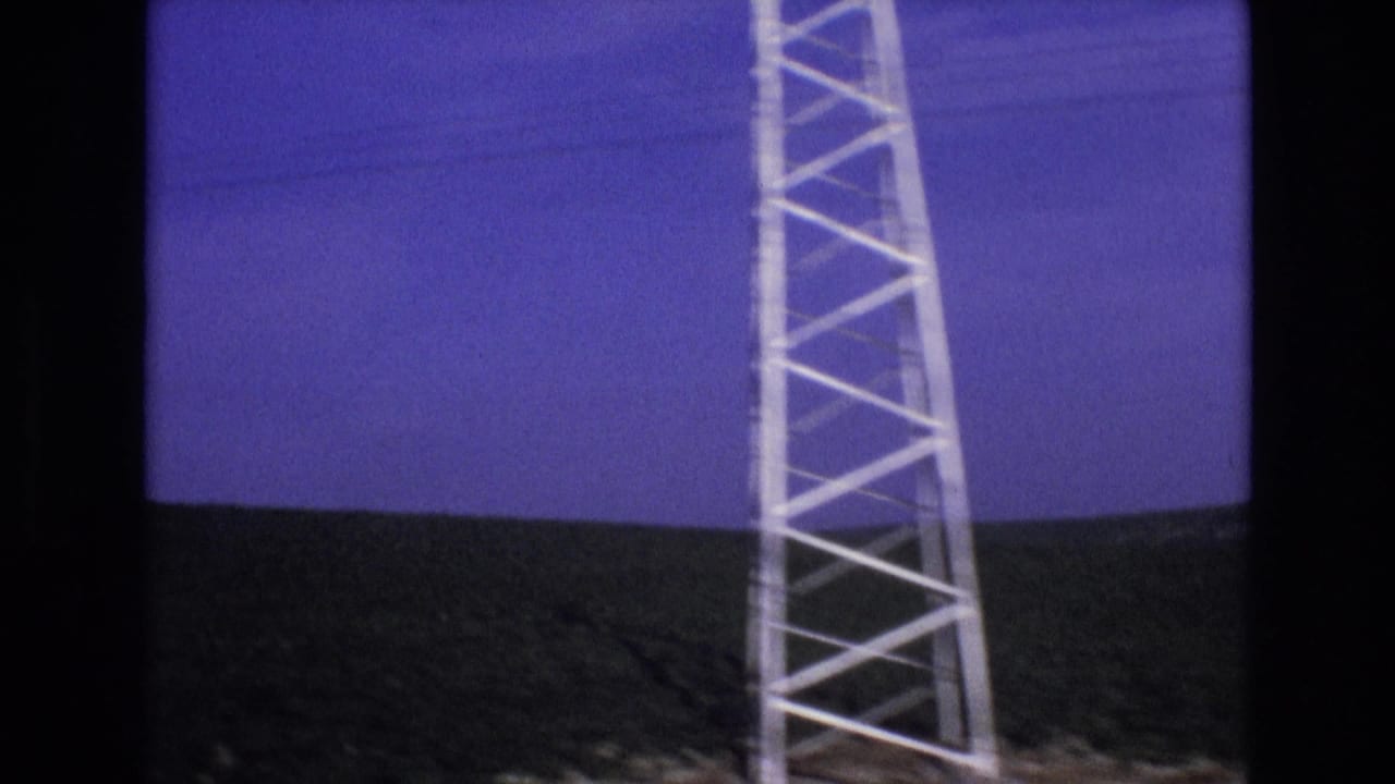 Roadside lattice tower beside hills and blue sky.