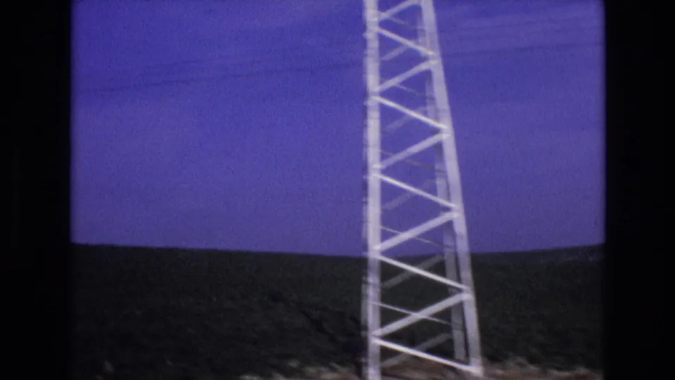 Metadata-supported 1972 Spain travel frame with a pale lattice tower at the roadside edge, low green hills, and a blue sky in soft 8mm color.
