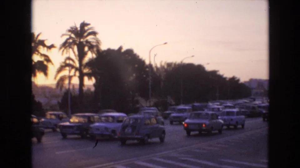 Metadata-supported 1972 Spain arrival frame showing a palm-lined town roadway with parked and moving cars, streetlights, and warm sunset light.