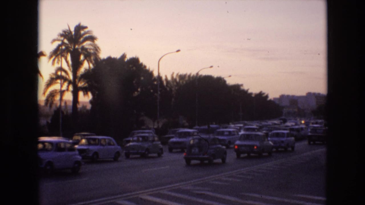 1972 Spain home-movie frame showing compact cars moving through dusk traffic on a broad road lined with palm trees and curved streetlights.