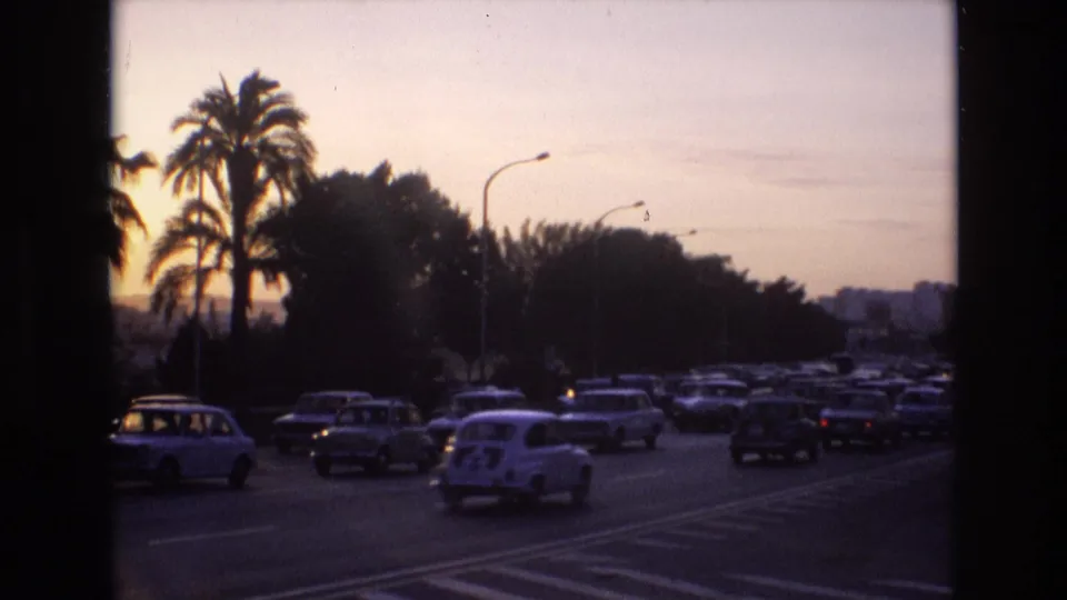 1972 Spain home-movie frame with compact cars spread across several lanes, a small white car in the foreground, silhouetted palms, and a fading evening sky.
