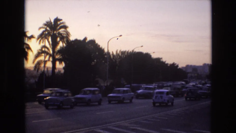 1972 Spain home-movie frame showing a stream of compact cars on a wide road at dusk, with palm trees and dark roadside foliage against a pale sky.