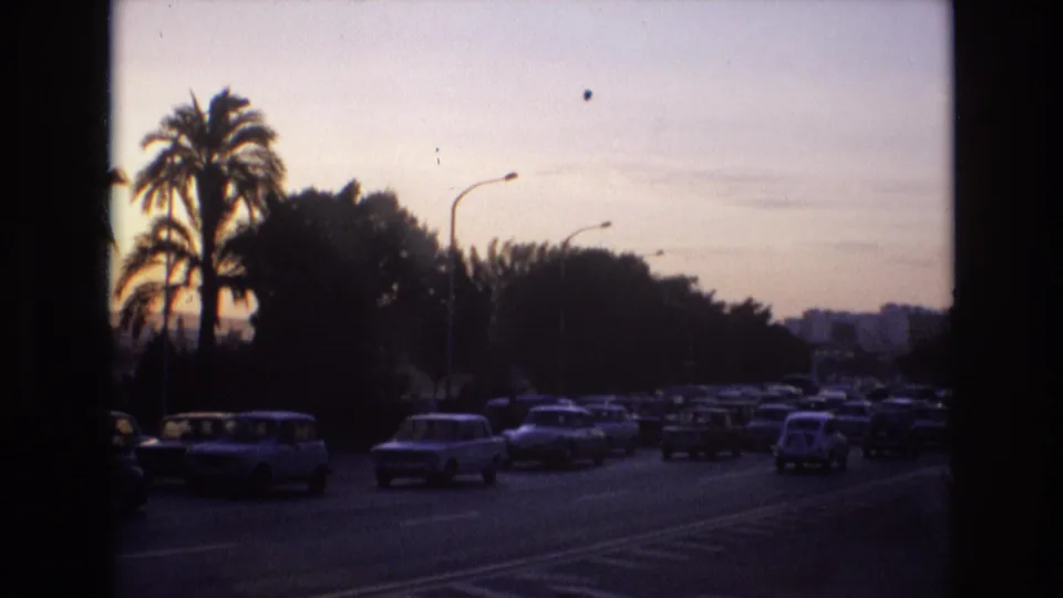 1972 Spain home-movie frame capturing dense dusk traffic on a multi-lane road, with silhouetted palms, curved streetlights, and distant buildings in soft focus.
