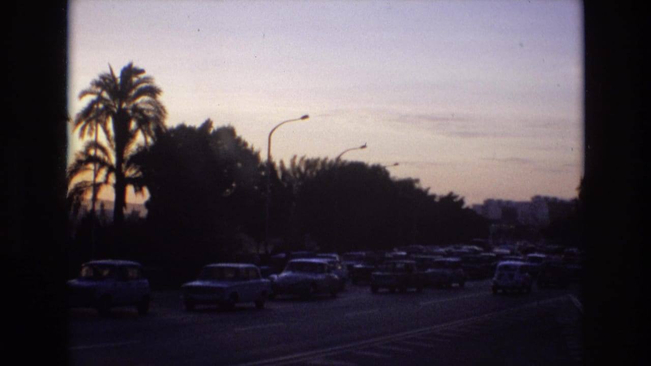 1972 Spain 8mm frame showing classic cars moving along a tree- and palm-lined road under a fading evening sky.