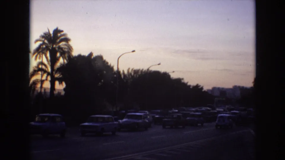 1972 Spain 8mm frame showing classic cars moving along a tree- and palm-lined road under a fading evening sky.