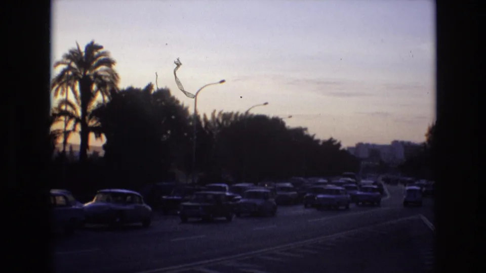 1972 Spain 8mm frame with busy classic-car traffic receding along a palm- and tree-lined roadway beneath a pale dusk sky.