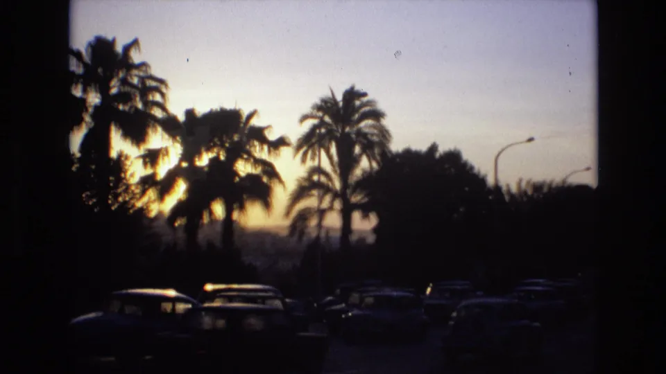 1972 Spain home-movie frame showing silhouetted cars and palm trees against a warm sunset sky on a busy road.