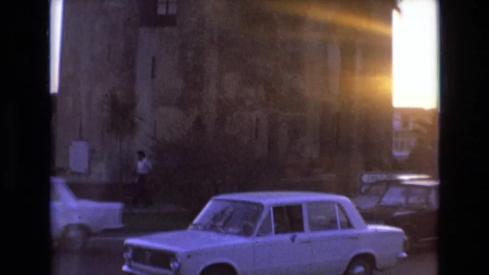 1972 Spain street view with a white sedan, weathered building wall, sunset flare, and a distant man walking along the sidewalk.