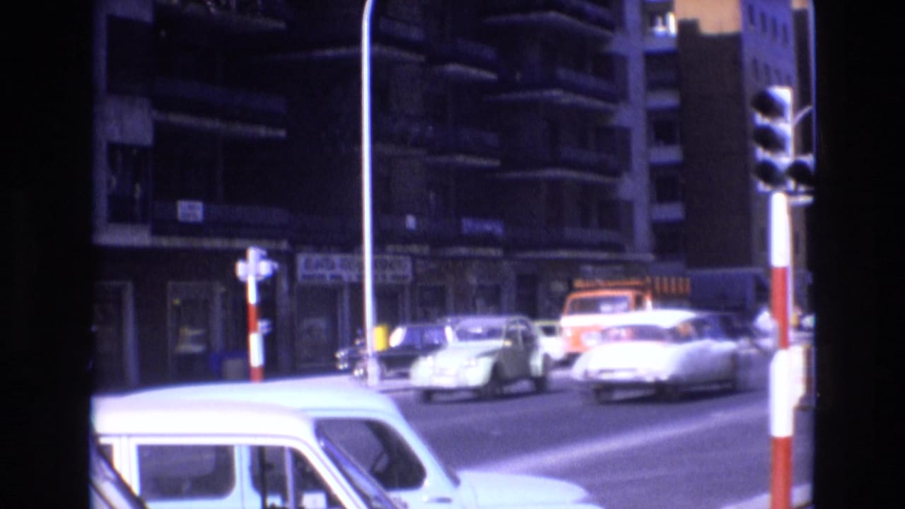 1972 Spain 8mm street view showing classic cars moving through an urban intersection, an orange truck near the curb, apartment balconies, storefront facades, and tall streetlights.