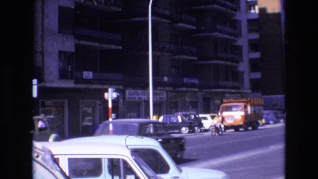 Cars and a motorcyclist move along a city street beside apartment buildings and an orange truck.