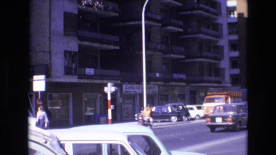 1972 Spain home-movie frame showing pedestrians near the sidewalk, parked and moving cars, an orange truck, multi-story apartment balconies, and a bright foreground car roof.