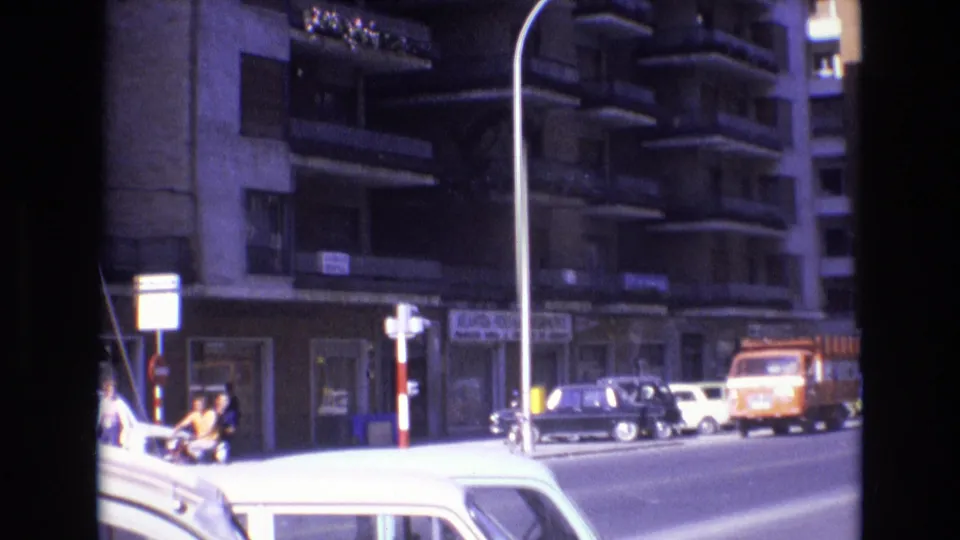 1972 Spain street view with pedestrians near the left sidewalk, a motorcyclist in traffic, parked cars, an orange truck, tall streetlights, and apartment blocks with balconies.