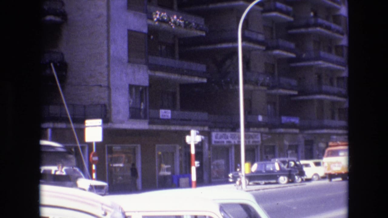 1972 Spain 8mm frame showing a multi-story apartment building with balconies, street signs, parked cars, and a tall curved streetlight in bright daylight.