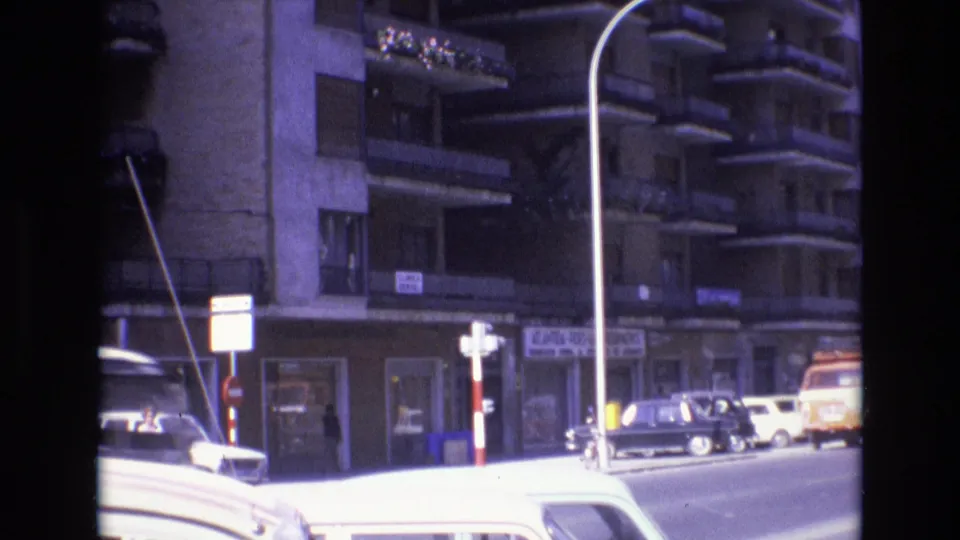 1972 Spain 8mm frame showing a multi-story apartment building with balconies, street signs, parked cars, and a tall curved streetlight in bright daylight.