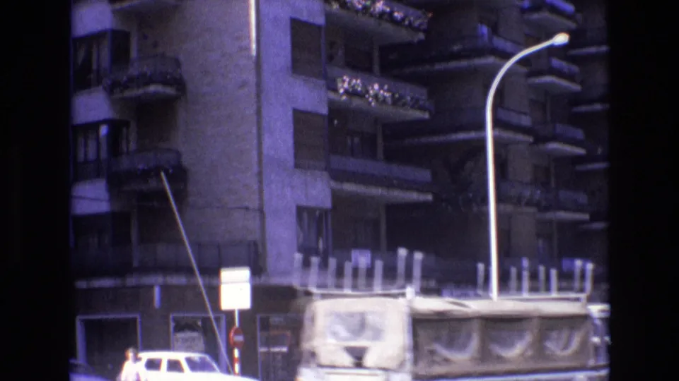 1972 Spain 8mm frame with a tall apartment facade, stacked balconies, a curved streetlight, a small pedestrian figure, and a covered truck or van moving through the street.