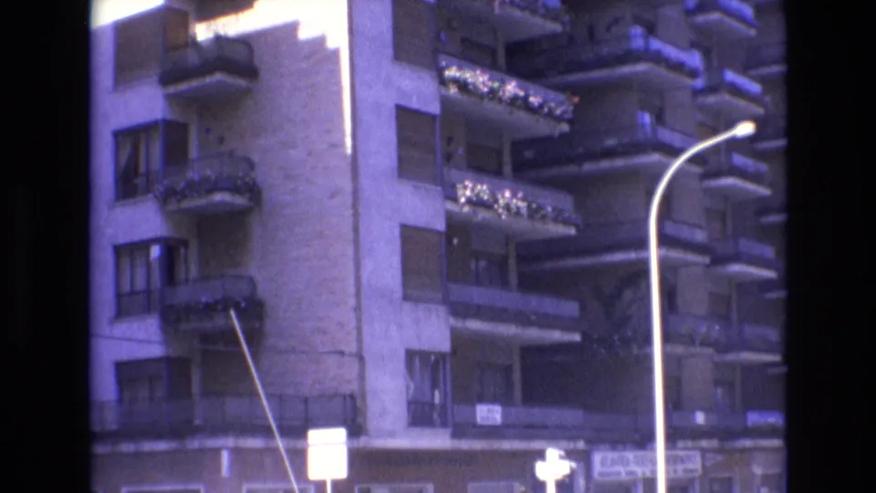 1972 Spain 8mm frame looking up at an apartment building with balconies and flower boxes, street signs near the ground floor, and a curved streetlight crossing the view.