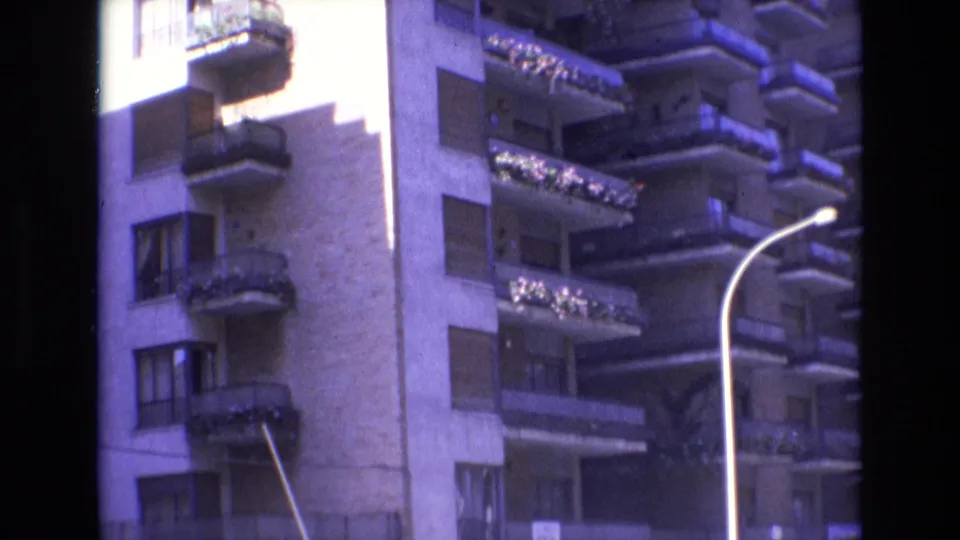 1972 Spain 8mm frame of a tall apartment building corner with shaded balconies, window shutters, flower boxes, and a prominent curved streetlight.