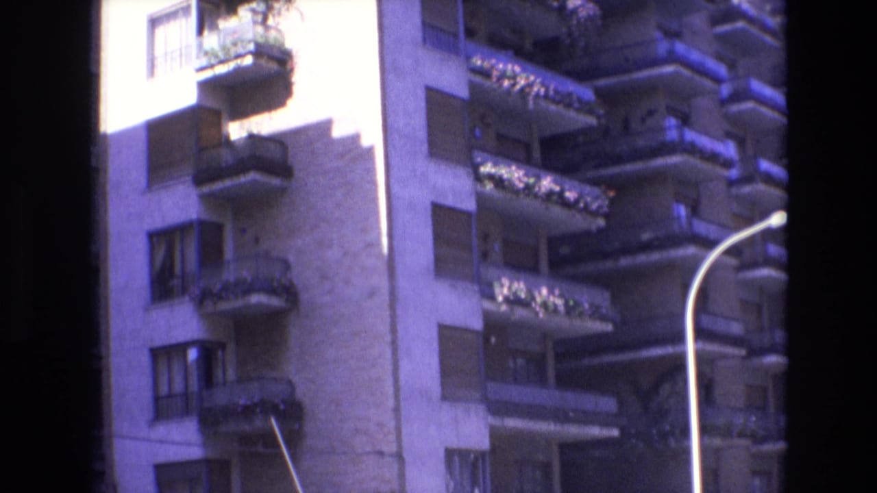 1972 Spain 8mm frame showing a sunlit apartment or resort building facade with planted balconies, shuttered windows, and a curved street lamp in front.
