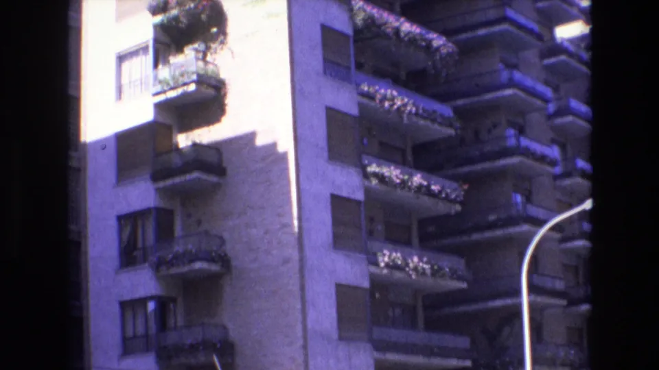 1972 Spain 8mm frame of a light-toned high-rise exterior with repeating balconies, planter boxes, shutters, and a partial curved lamp in the foreground.