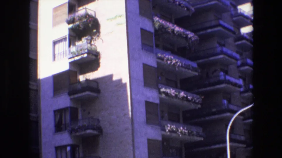 1972 Spain 8mm frame with a closer street-level view of planted balconies and windows on a sunlit high-rise exterior, with darker neighboring balconies beside it.