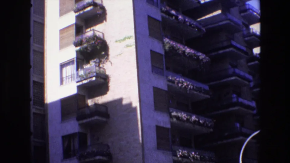1972 Spain 8mm frame showing the same high-rise exterior with planted balconies, window shutters, strong sun and shadow contrast, and a curved street lamp at frame right.