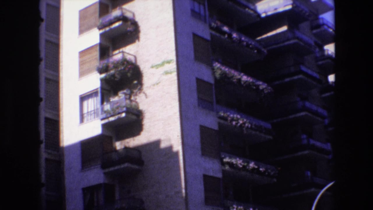 1972 Spain 8mm frame showing a pale multi-story apartment facade with projecting balconies, dark railings, shuttered windows, plants, and strong sunlight.