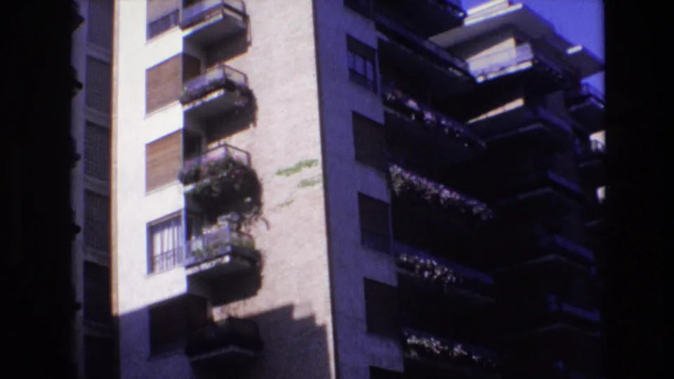 1972 Spain 8mm frame of a residential building exterior with repeated balconies, window shutters, a pale wall surface, and heavy shadow across part of the facade.