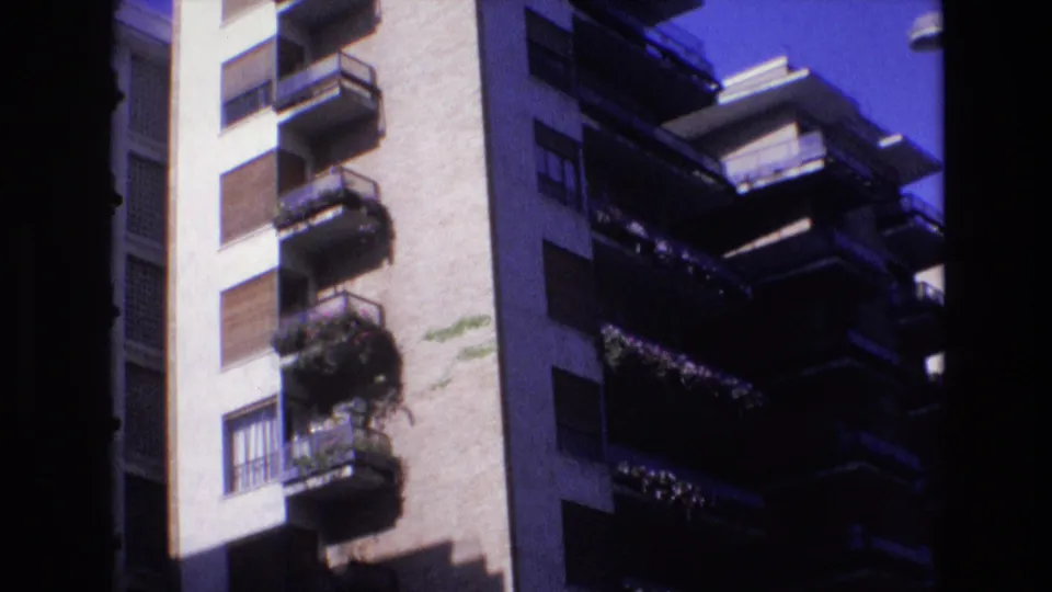 1972 Spain 8mm frame showing the building facade from a lower angle, with stacked balconies, dark railings, potted greenery, shuttered windows, and sunlit plaster.