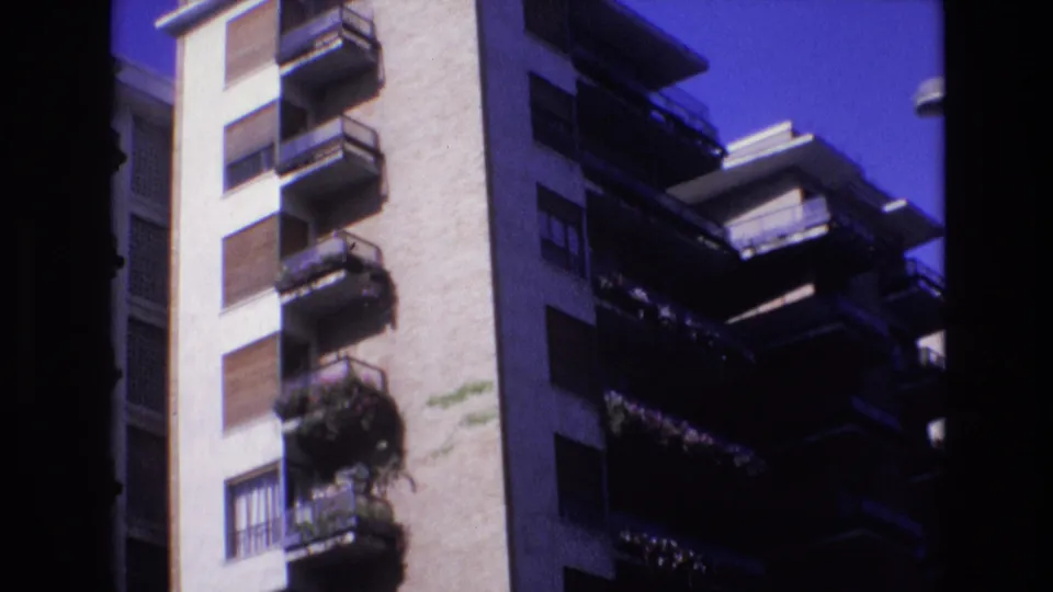 1972 Spain 8mm frame looking upward at upper-floor apartment balconies, shuttered windows, a pale facade, deep shadows, and clear blue sky.