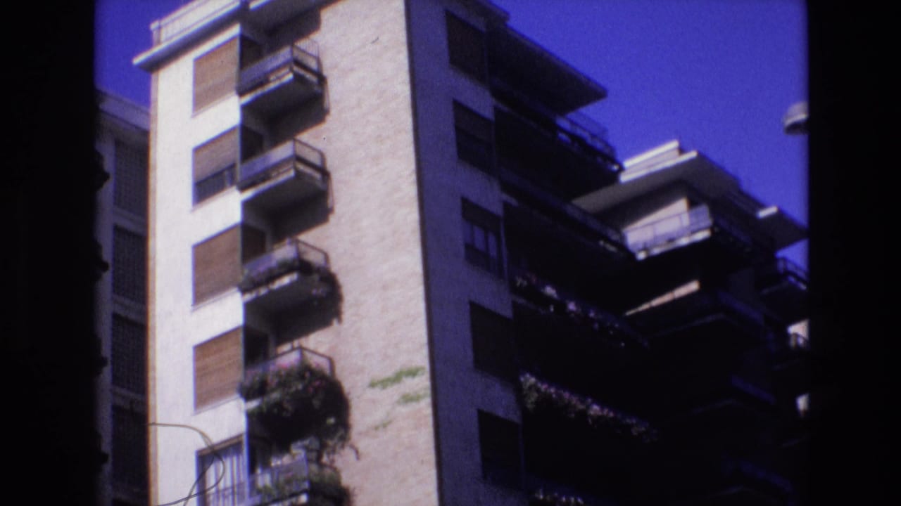 1972 Spain home-movie frame showing a tall light-colored apartment facade with stacked balconies, closed shutters, potted plants, and a deep blue sky.