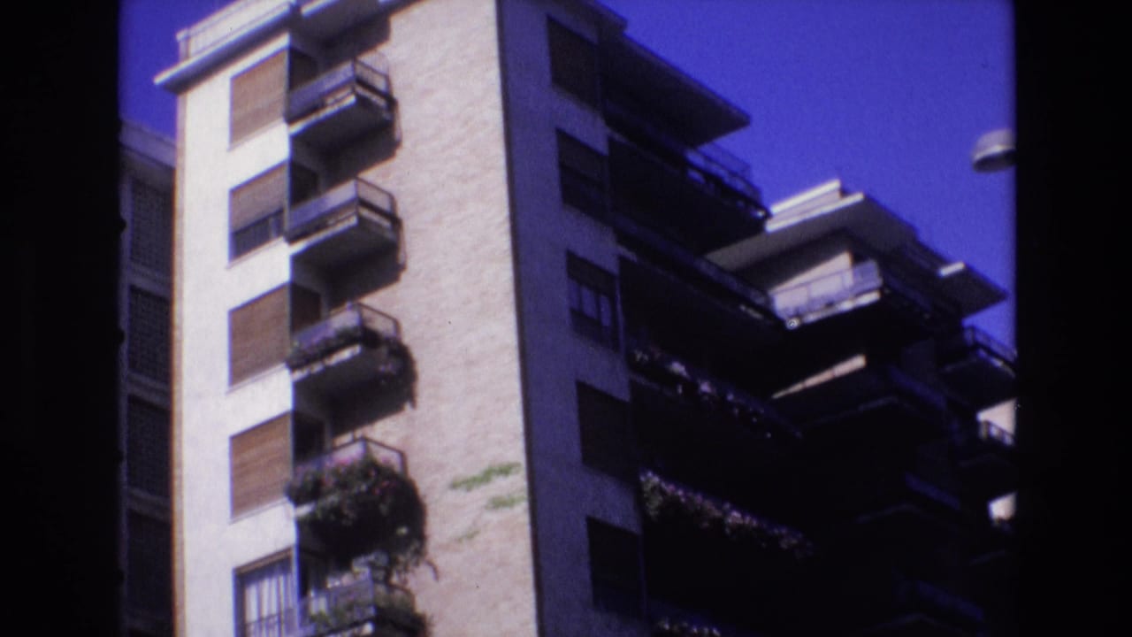 Tall apartment facade with balcony railings, shutters, and plants in bright sun.
