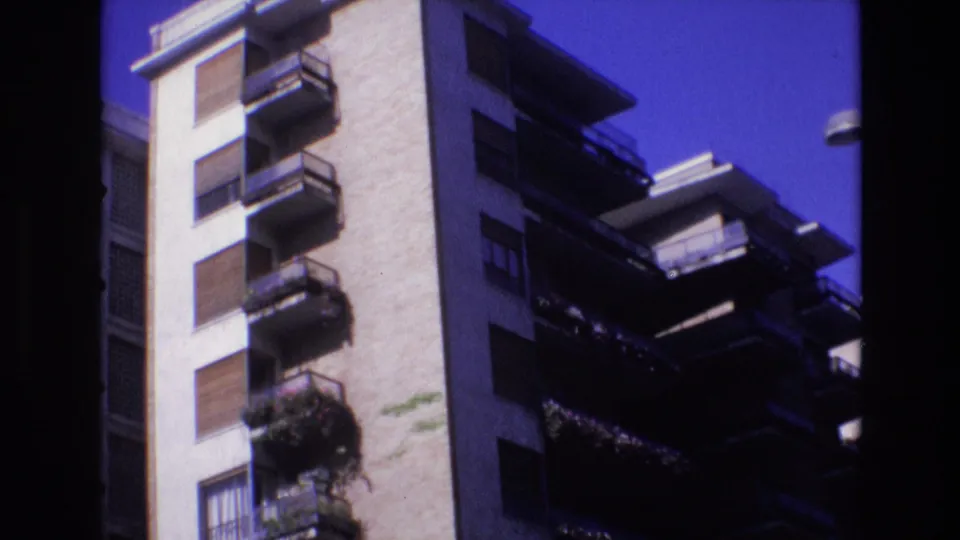 1972 Spain frame of a tall residential facade with sunlit wall sections, dark balcony shadows, shutters, railing lines, and balcony greenery.