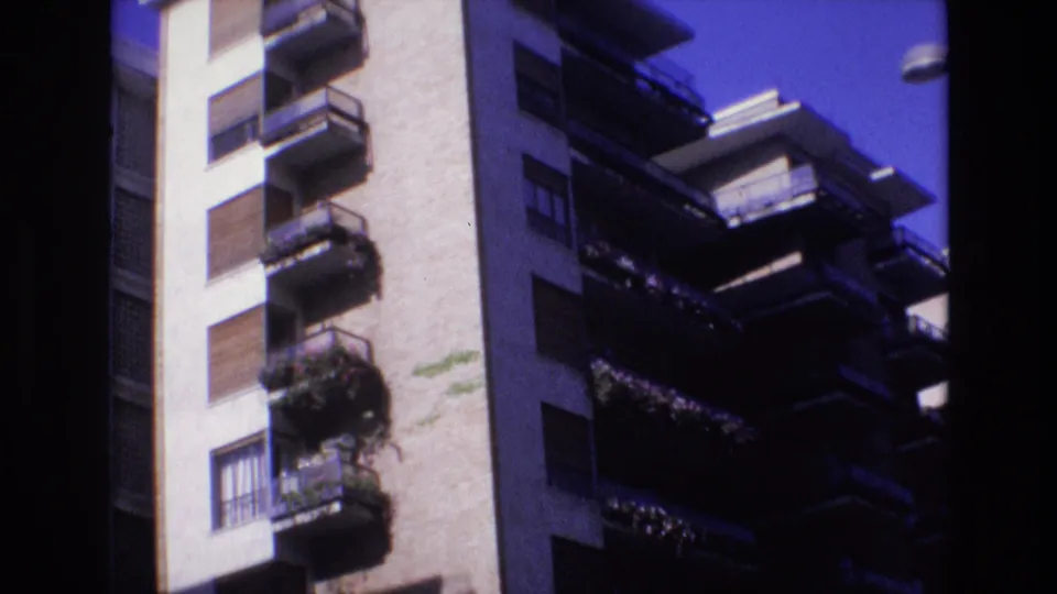 1972 Spain home-movie still showing multiple apartment balconies with railings and plants, a pale facade, shuttered windows, and dark edges from the film frame.
