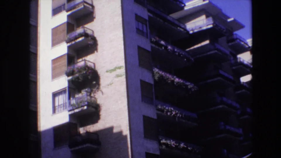 1972 Spain frame with a closer view of the apartment building's balcony stack, showing potted plants, dark railings, shuttered windows, and heavy shadow across the facade.