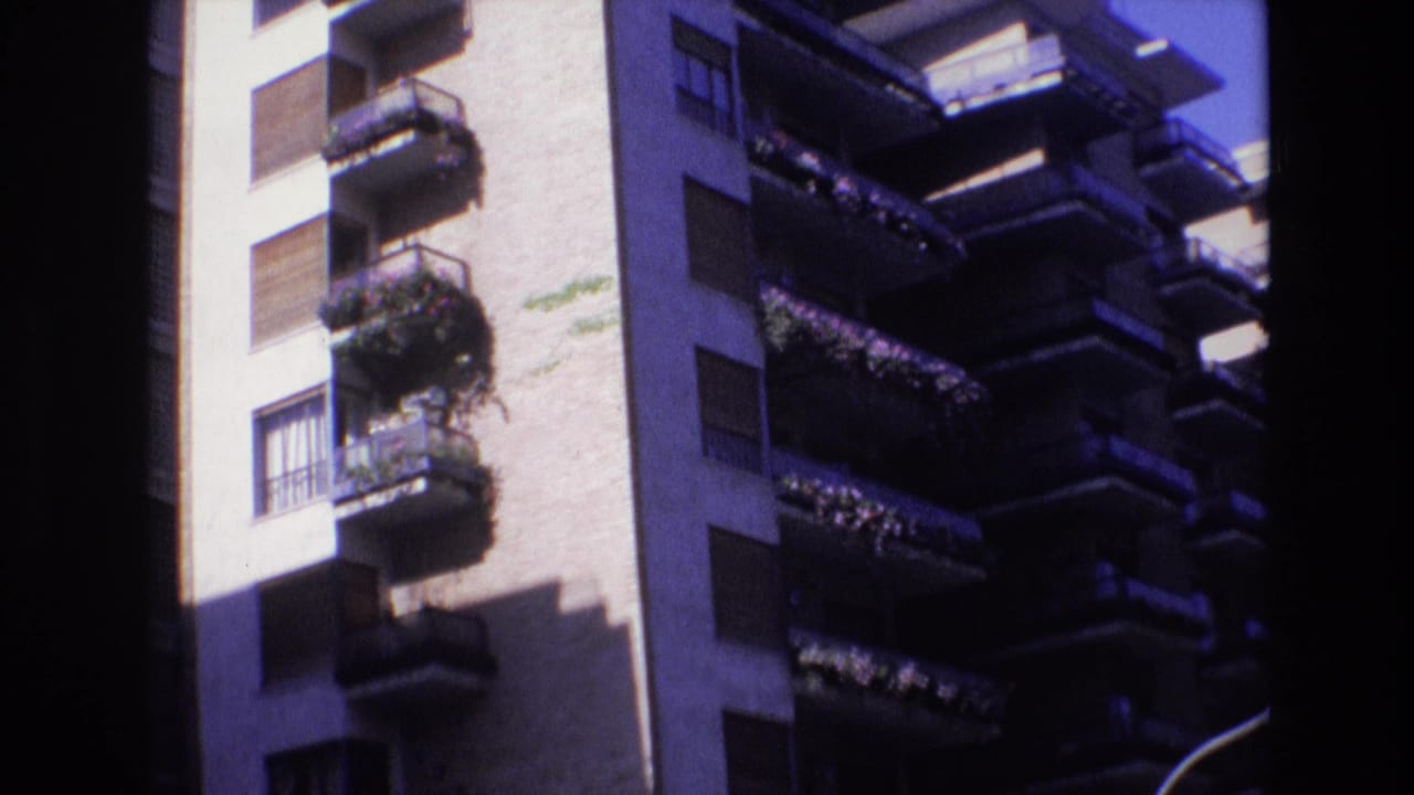 1972 Spain home-movie frame showing a sunlit apartment building facade with stacked balconies, shutters, plants, and strong shadow across the wall.