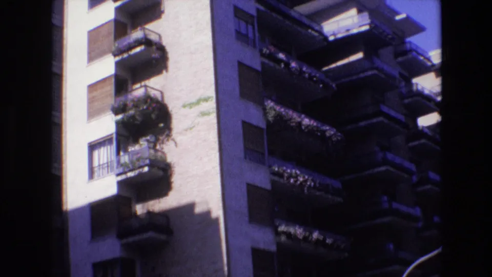 1972 Spain home-movie frame showing a sunlit apartment building facade with stacked balconies, shutters, plants, and strong shadow across the wall.