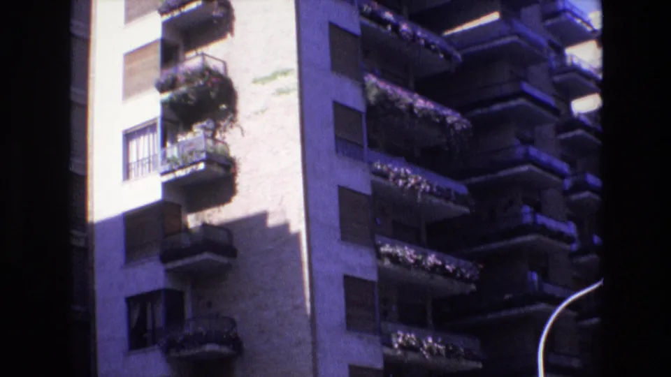 1972 Spain frame with the apartment block seen through a home-movie scan, showing balcony rows, shuttered windows, planted railings, and a dark vertical frame edge.
