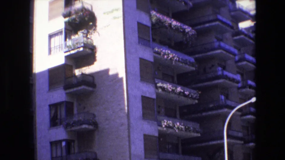 1972 Spain home-movie frame showing a closer street-level view of an apartment facade with planted balconies, rectangular windows, hard sun, and a curved streetlight at the edge.