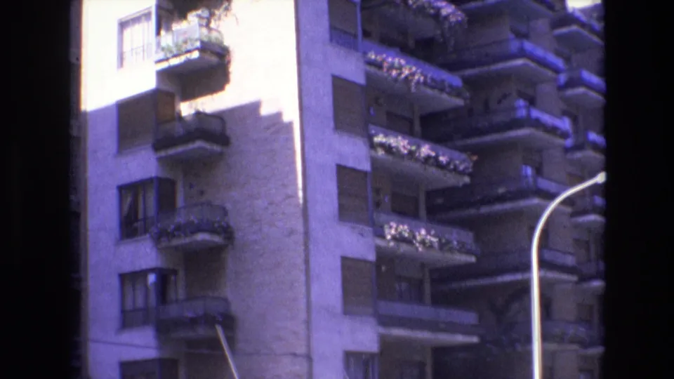1972 Spain frame showing a tall residential building with repeating balconies, window shutters, balcony greenery, strong afternoon-like light, and dark film borders.