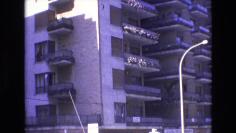 Spain, 1972, with a slightly shifted view of the apartment building, balconies with plants, a prominent curved streetlamp, and street-level commercial frontage.