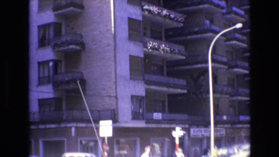 Spain, 1972, showing the lower floors of an apartment building with balconies, street signs, shop entrances, and a partially visible bright vehicle passing through the frame.
