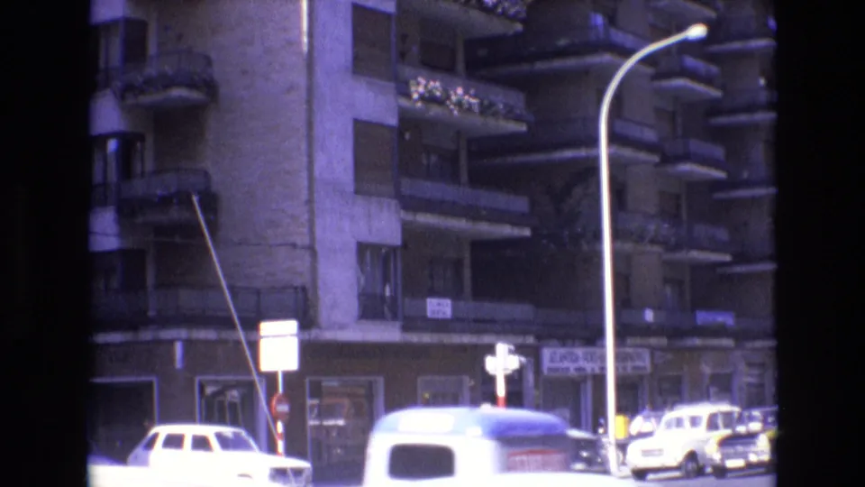 Spain, 1972, with a white van and other cars near a crossing below an apartment building with balconies, street signs, shopfronts, and a curved streetlamp.