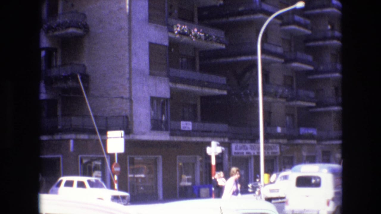 1972 Spain home-movie frame showing a sunlit urban street corner with apartment facades, a white car, a light-colored van, traffic signs, and pedestrians near the curb.