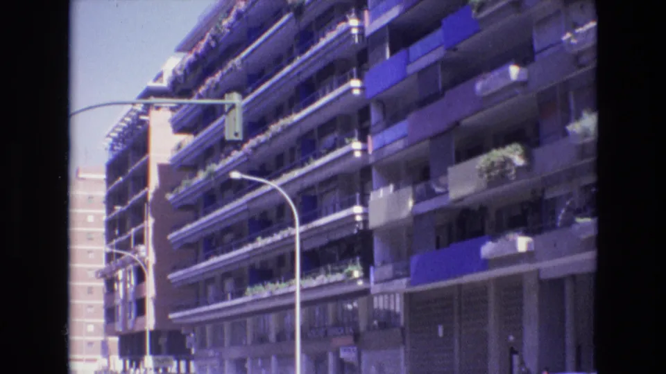 1972 Spain home-movie frame looking up a residential city street toward apartment balconies with blue awnings, balcony plants, curved streetlights, and a suspended traffic signal.