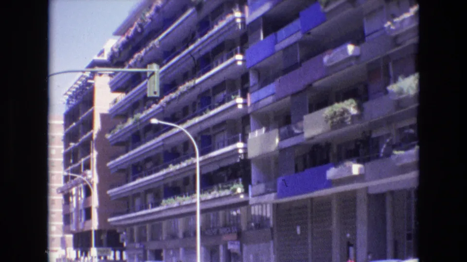 1972 Spain home-movie frame showing a closer street-level view of a multi-story apartment facade with repeating balconies, blue awnings, plants, and curved streetlights.