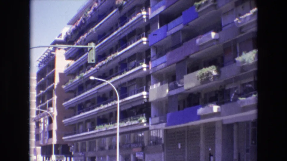 1972 Spain home-movie frame of a sunlit apartment block with blue awnings, balcony plants, curved streetlights, and a traffic signal above the street.