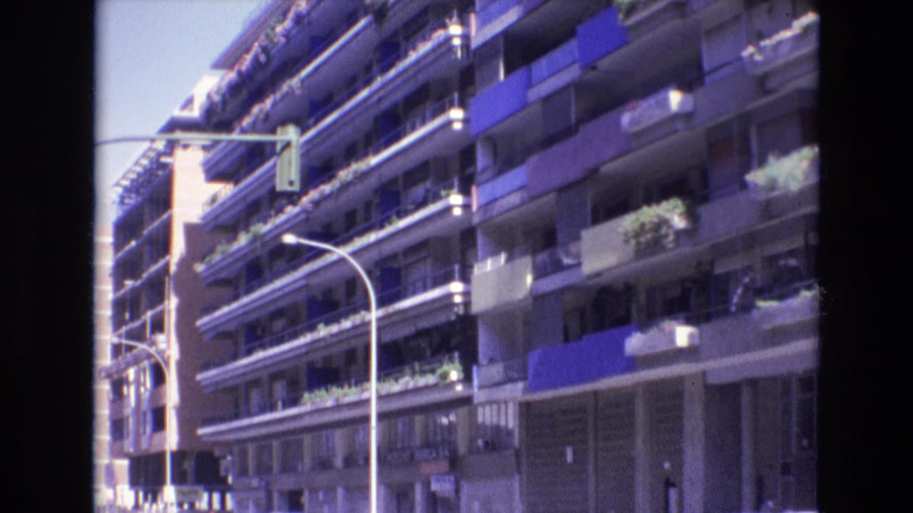 1972 Spain home-movie frame showing a sunlit multi-story apartment facade with rows of balconies, blue awnings, planted ledges, a street lamp, and a traffic signal.