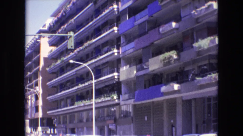 1972 Spain street-level view of a large apartment building with repeating balcony lines, blue awnings, planted balcony boxes, a curved lamp post, and a suspended traffic light.