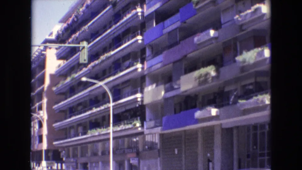 1972 Spain frame with a slightly closer home-movie view of stacked apartment balconies, blue awnings, planted ledges, a street lamp, and a traffic signal in front of the building.